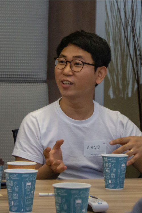 Man in white t-shirt at a table talking to someone off screen, blue paper cups on table.